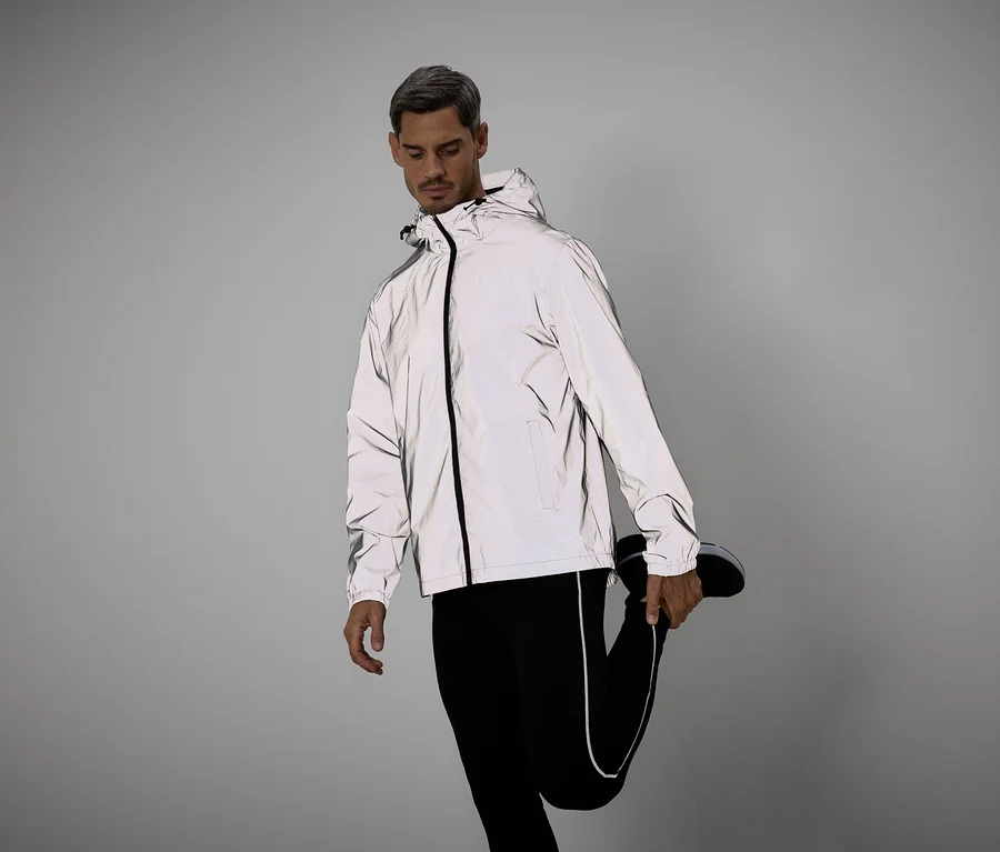 Mann in reflektierender Jacke und Leggings dehnt Bein. Er trägt schwarze Schuhe vor grauem Hintergrund.