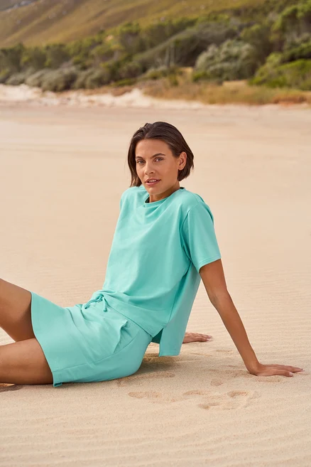 Frau sitzt am Strand und trägt ein hellblaues T-Shirt und Shorts.
