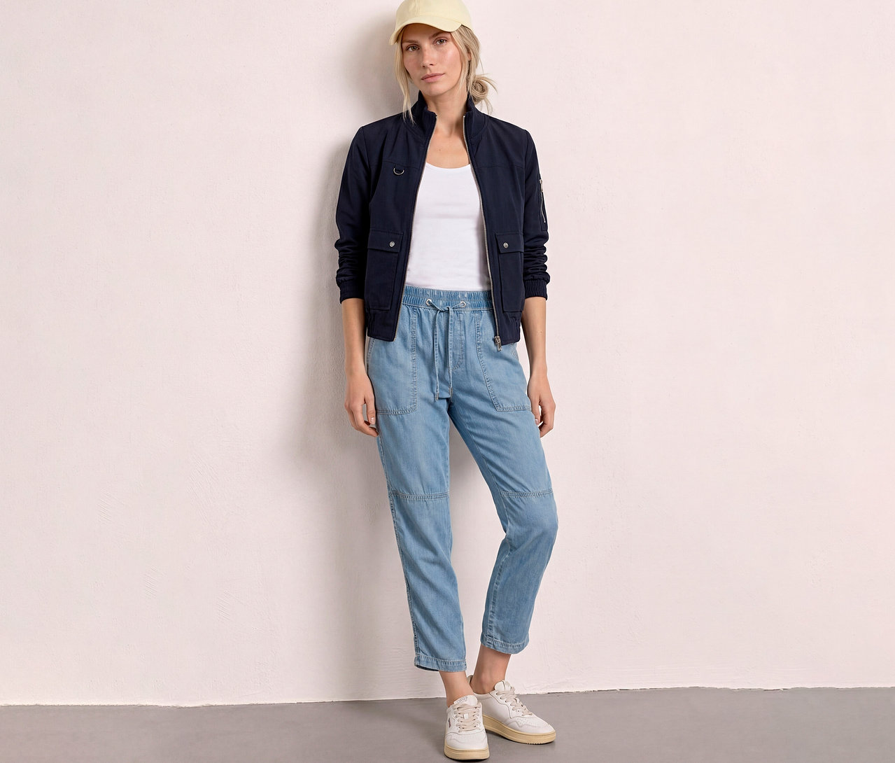 Frau in marineblauer Jacke, weißem Top, hellblauen Jeans und gelber Kappe steht vor einer Wand.