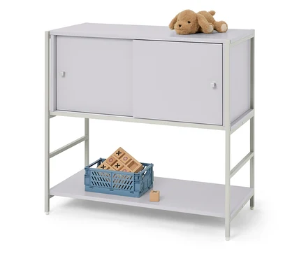 Ein beiger Stoffhund liegt auf einem grauen Schrank mit Schiebetüren. Unter dem Schrank steht eine blaue Kiste mit Holzspielzeug.