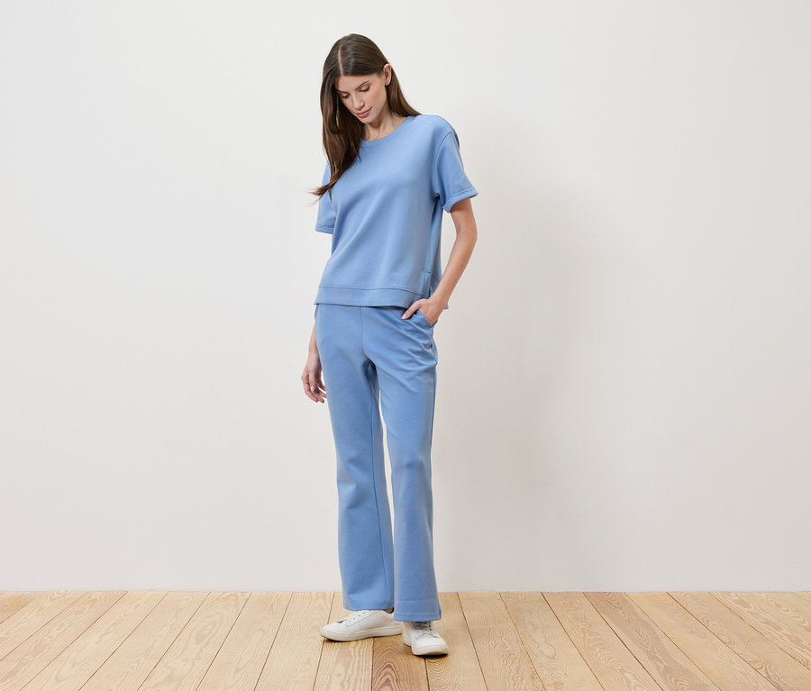 Frau mit braunen Haaren trägt ein hellblaues T-Shirt und passende Hosen und hat die Hand in der Hosentasche.