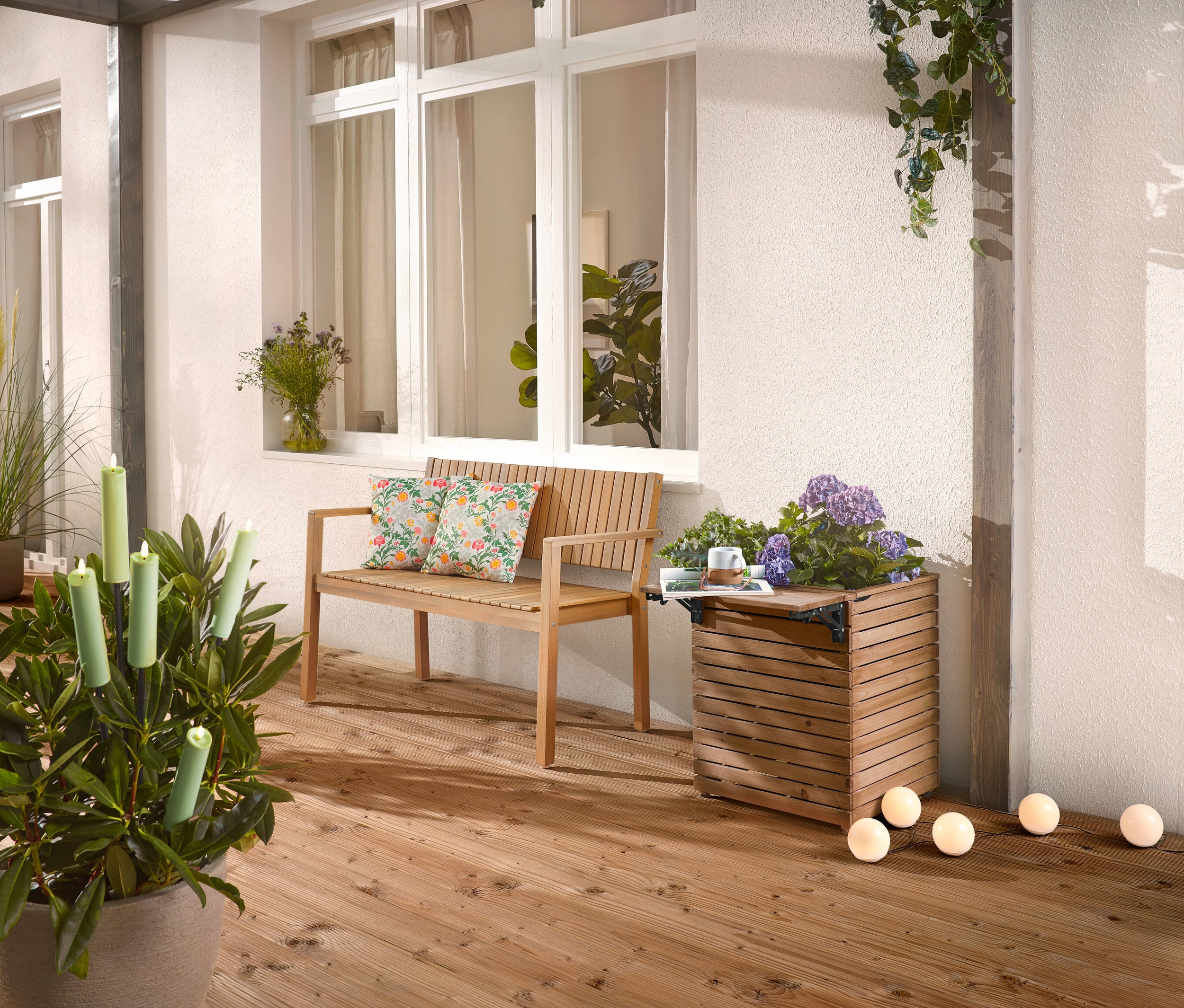 Auf einer Holzterrasse stehen eine Holzbank mit Kissen, ein Pflanzkasten mit Blumen und eine Topfpflanze mit Kerzen.