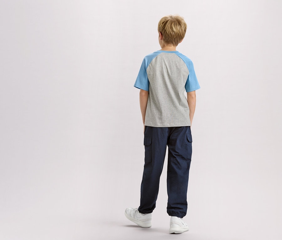 Rückansicht eines Kindes mit blonden Haaren, das ein graues T-Shirt mit blauen Ärmeln, eine marineblaue Hose und weiße Turnschuhe trägt.