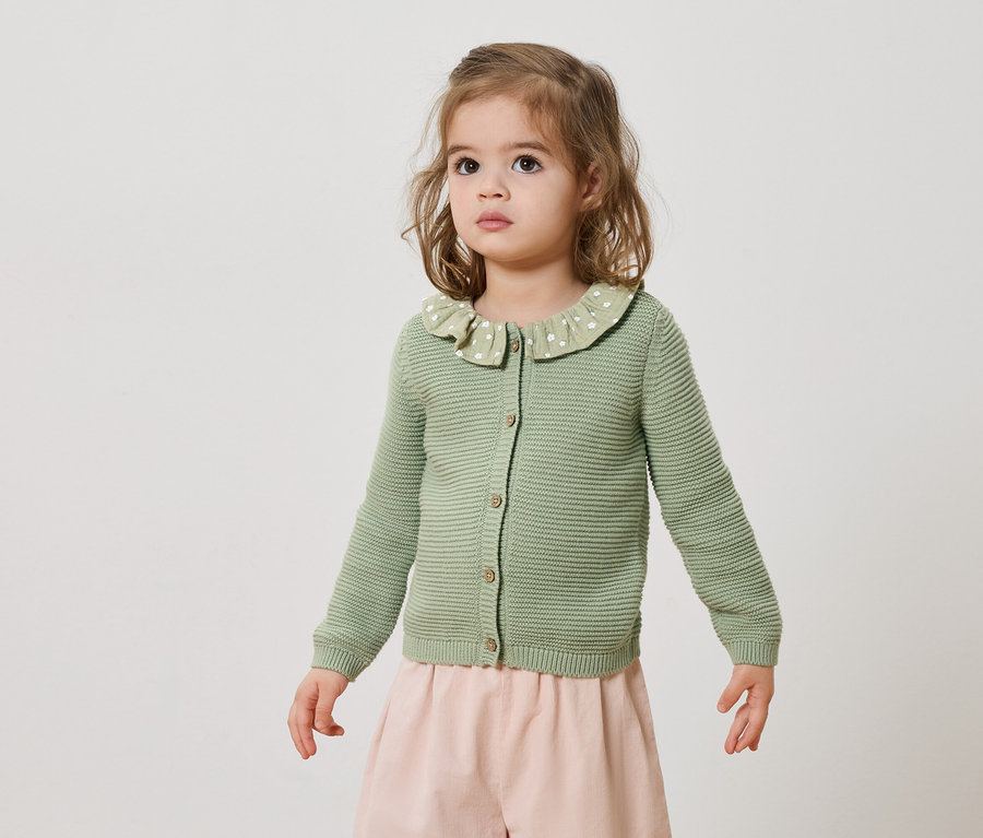 Ein kleines Mädchen trägt eine grüne Strickjacke mit Knopfleiste und einen rosa Rock.