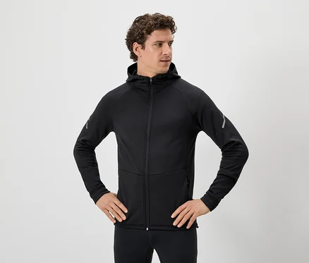 Mann mit lockigem Haar, schwarzer Kapuzenjacke mit Reißverschluss und schwarzen Leggings, die Hände in den Hüften.