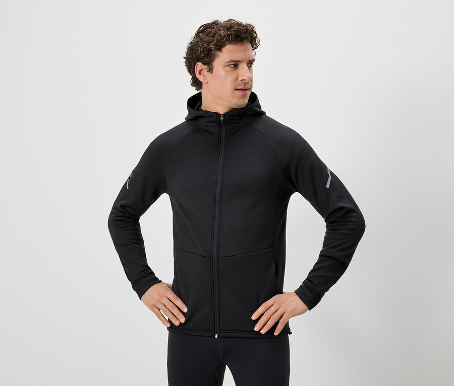 Mann mit lockigem Haar, schwarzer Kapuzenjacke mit Reißverschluss und schwarzen Leggings, die Hände in den Hüften.