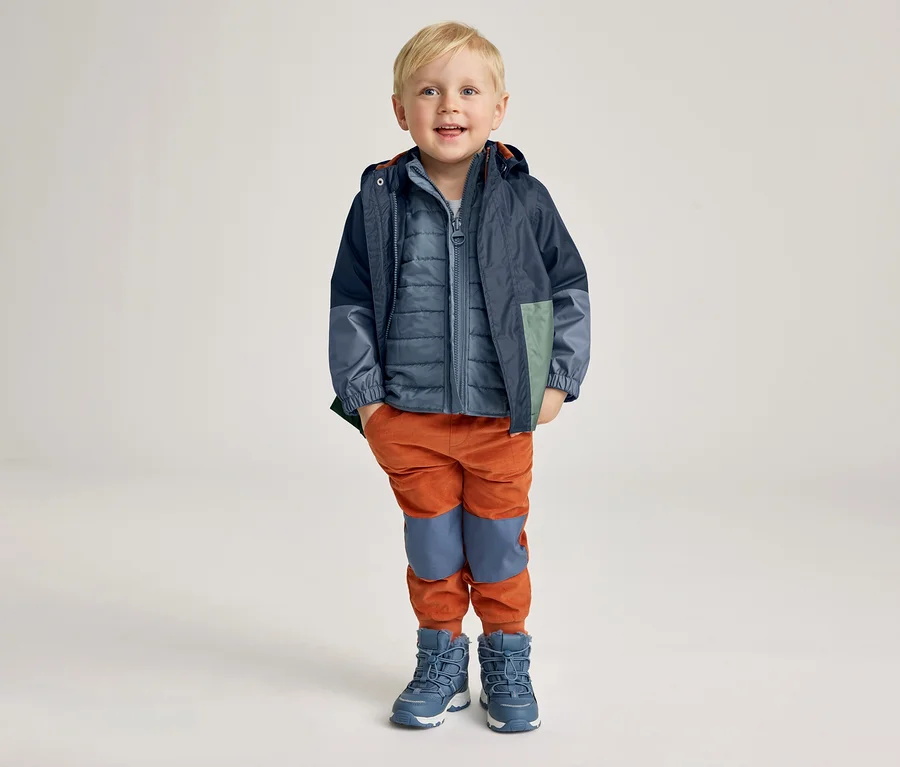 Ein lächelnder Junge mit blonder Kurzhaarfrisur trägt eine blaue Jacke, eine blaue Steppweste, orangefarbene Hosen und blaue Stiefel.