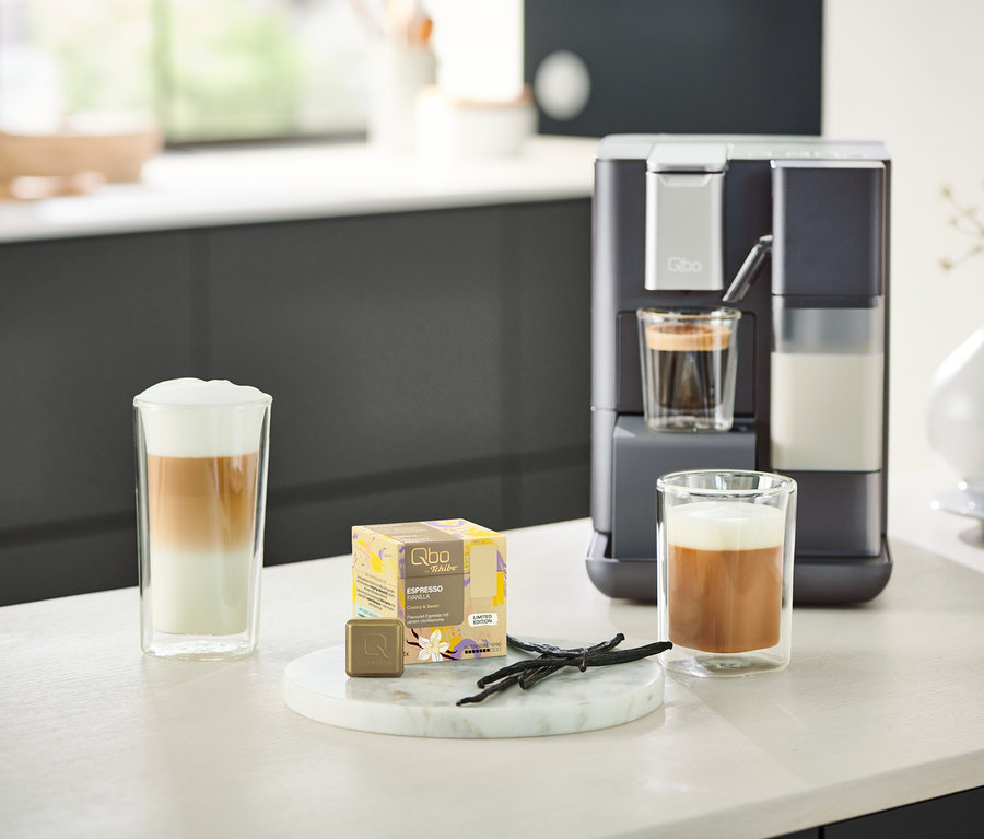 Ein Qbo MILK MASTER Gentle Grey steht neben zwei Gläsern Kaffee. Eine rote Qbo MILK MASTER ist im Hintergrund sichtbar.