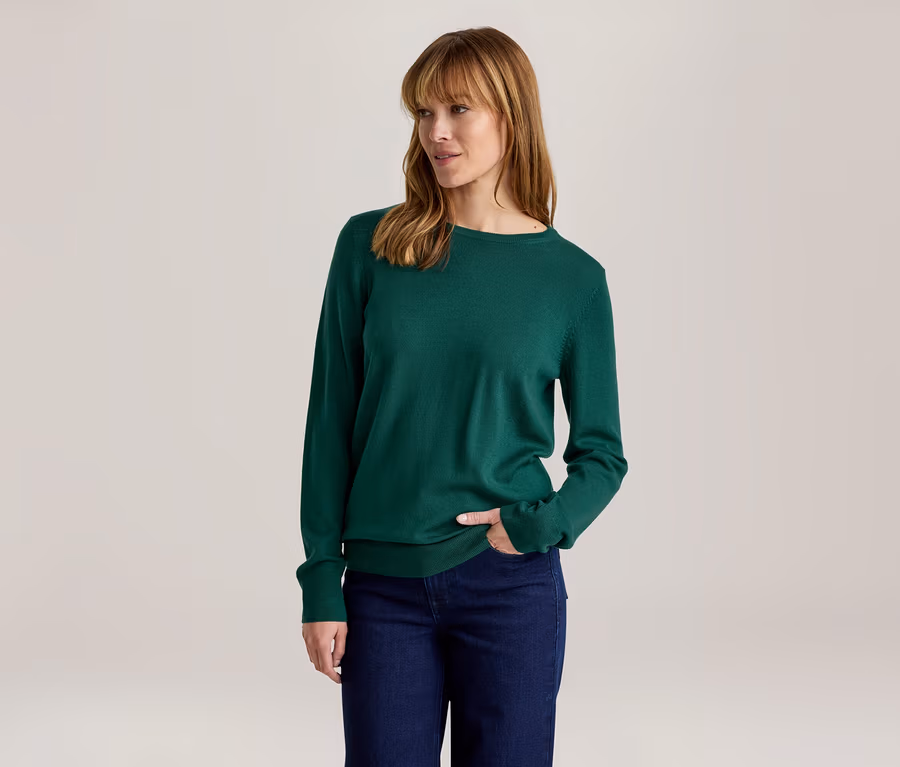 Frau in einem grünen Langarmshirt und dunklen Jeans, mit der rechten Hand in der Tasche, blickt nach rechts.