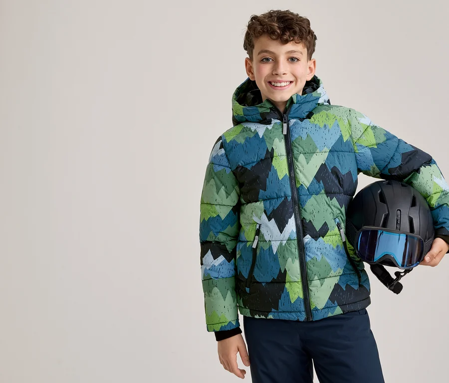 Ein lächelnder Junge in einer grün, blau und schwarz gemusterten Winterjacke hält einen schwarzen Skihelm mit blauer Skibrille.