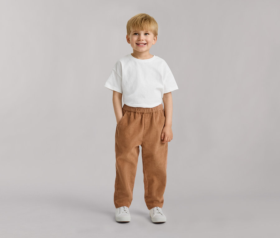 Junger Junge mit blondem Haar, lächelnd, trägt ein weißes T-Shirt, braune Cordhose und weiße Turnschuhe, Hände in den Taschen.