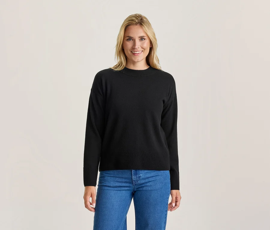 Lächelnde blonde Frau in einem schwarzen Langarmpullover und blauen Jeans vor hellem Hintergrund.