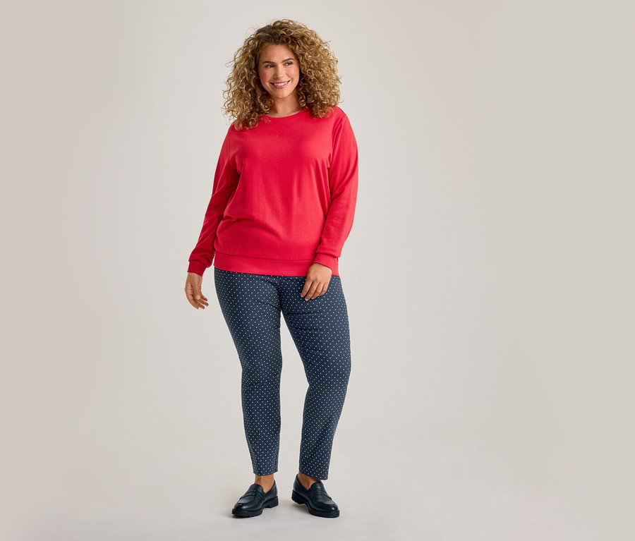 Lächelnde Frau mit lockigem Haar, die ein rotes Langarmshirt, dunkelblaue Hosen mit weißen Punkten und schwarze Schuhe trägt.