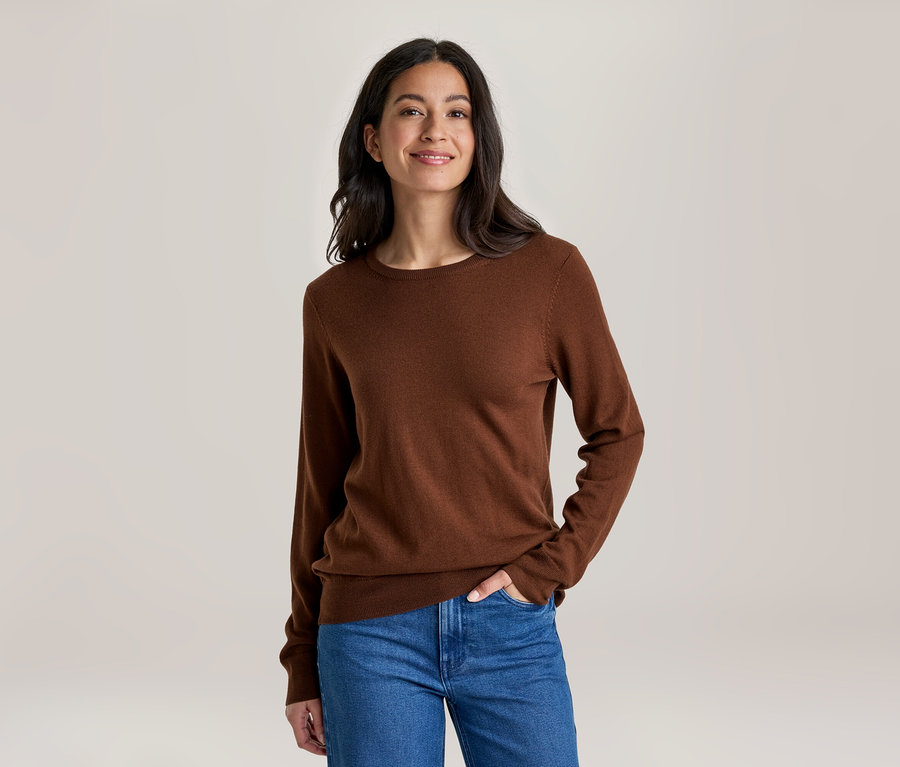 Eine lächelnde Frau mit dunklen Haaren trägt ein braunes Langarmshirt und blaue Jeans, eine Hand in der Tasche, vor hellem Hintergrund.