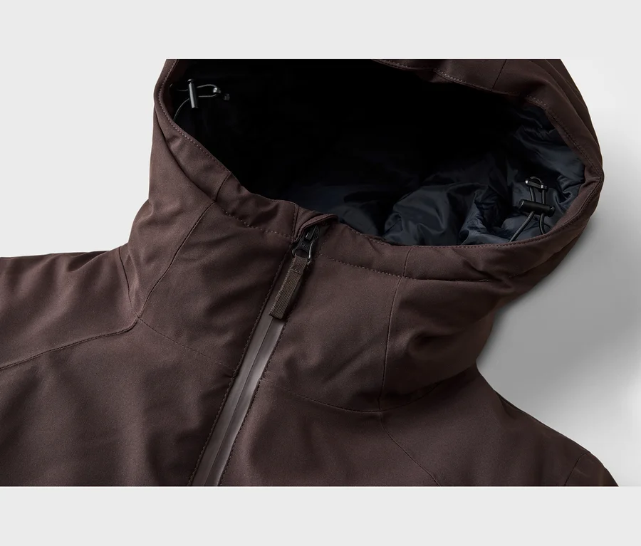 Nahaufnahme einer braunen Jacke mit teilweise geöffnetem Reißverschluss, der ein dunkles Innenfutter zeigt. Die Kapuze ist aufgesetzt und zeigt einen schwarzen Kordelzug mit Stopper.