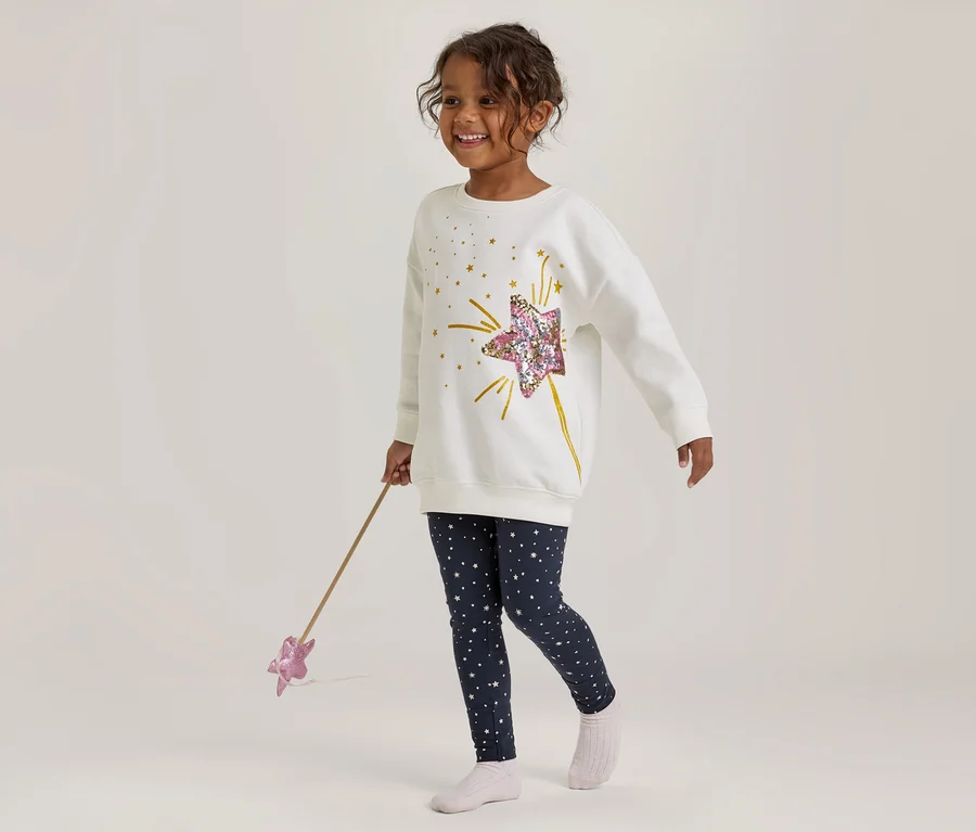 Lächelndes Mädchen in weißem Langarmshirt mit Pailletten-Stern und dunkelblauen Leggings mit Sternen, das einen rosa Sternenzauberstab hält.