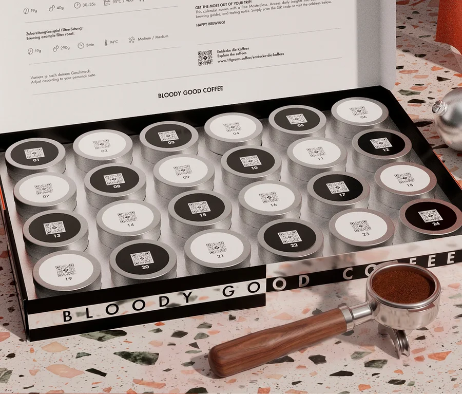 Eine offene Kaffee-Adventskalenderbox mit der Aufschrift „BLOODY GOOD COFFEE“ enthält 24 nummerierte silberne und schwarze Kaffeekapseln. Daneben liegen ein Siebträger mit Holzgriff, gefüllt mit gemahlenem Kaffee, und zwei silberne Weihnachtskugeln auf einer gesprenkelten Arbeitsplatte.