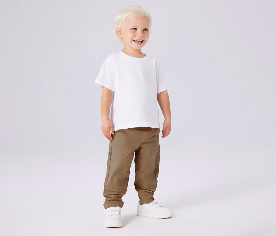 Lächelnder Junge mit weißem T-Shirt und NAME IT Kinder-Twillhose in Khaki und weißen Schuhen.