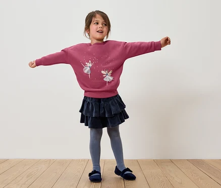Mädchen steht mit ausgestreckten Armen, trägt ein rosa Sweatshirt mit Ballerinas, einen dunkelblauen Rock mit Rüschen, graue Strumpfhosen und blaue Schuhe.