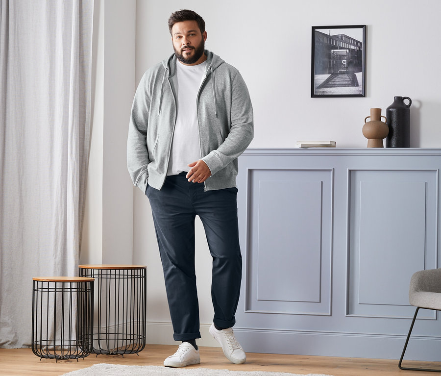Ein Mann steht und trägt eine graue Sweatjacke, ein weißes T-Shirt, blaue Chinos und weiße Turnschuhe. Ebenfalls abgebildet sind 4 Paar Merino-Socken, schwarz, dunkelblau, anthrazit und grau.
