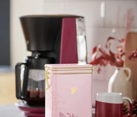 Eine rosa Kaffeedose steht auf einer Küchentheke neben einer Kaffeemaschine und einer rot-weißen Tasse.