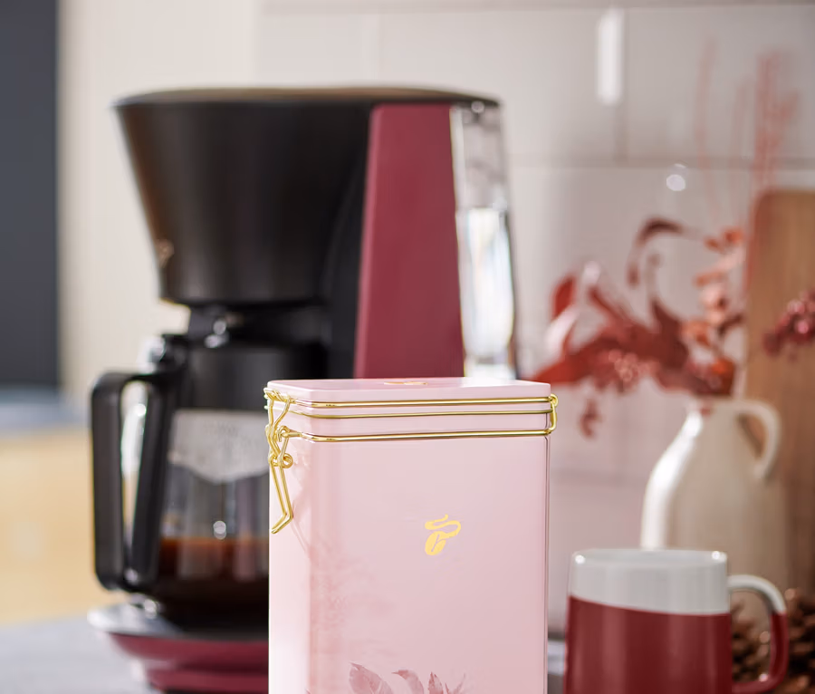 Eine rosa Kaffeedose steht auf einer Küchentheke neben einer Kaffeemaschine und einer rot-weißen Tasse.