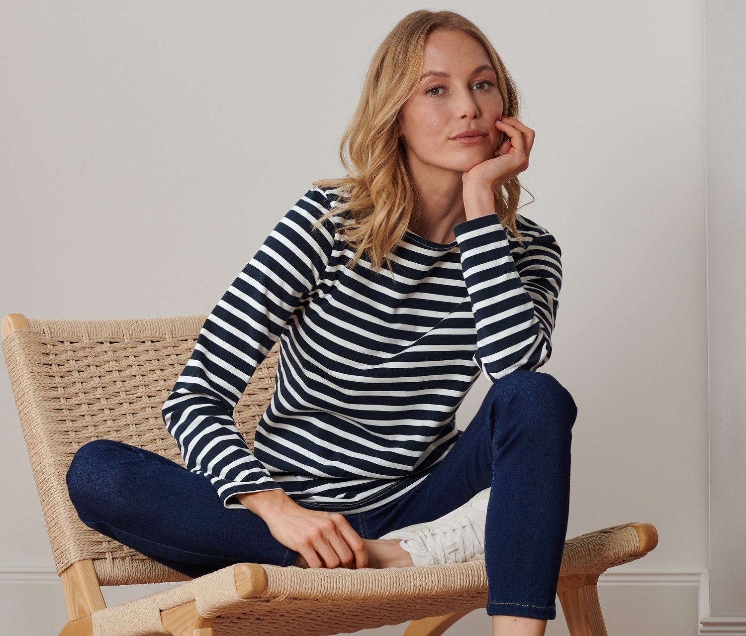 Blonde Frau sitzt in einem Stuhl und trägt ein dunkelblau-cremeweiß gestreiftes Langarmshirt.
