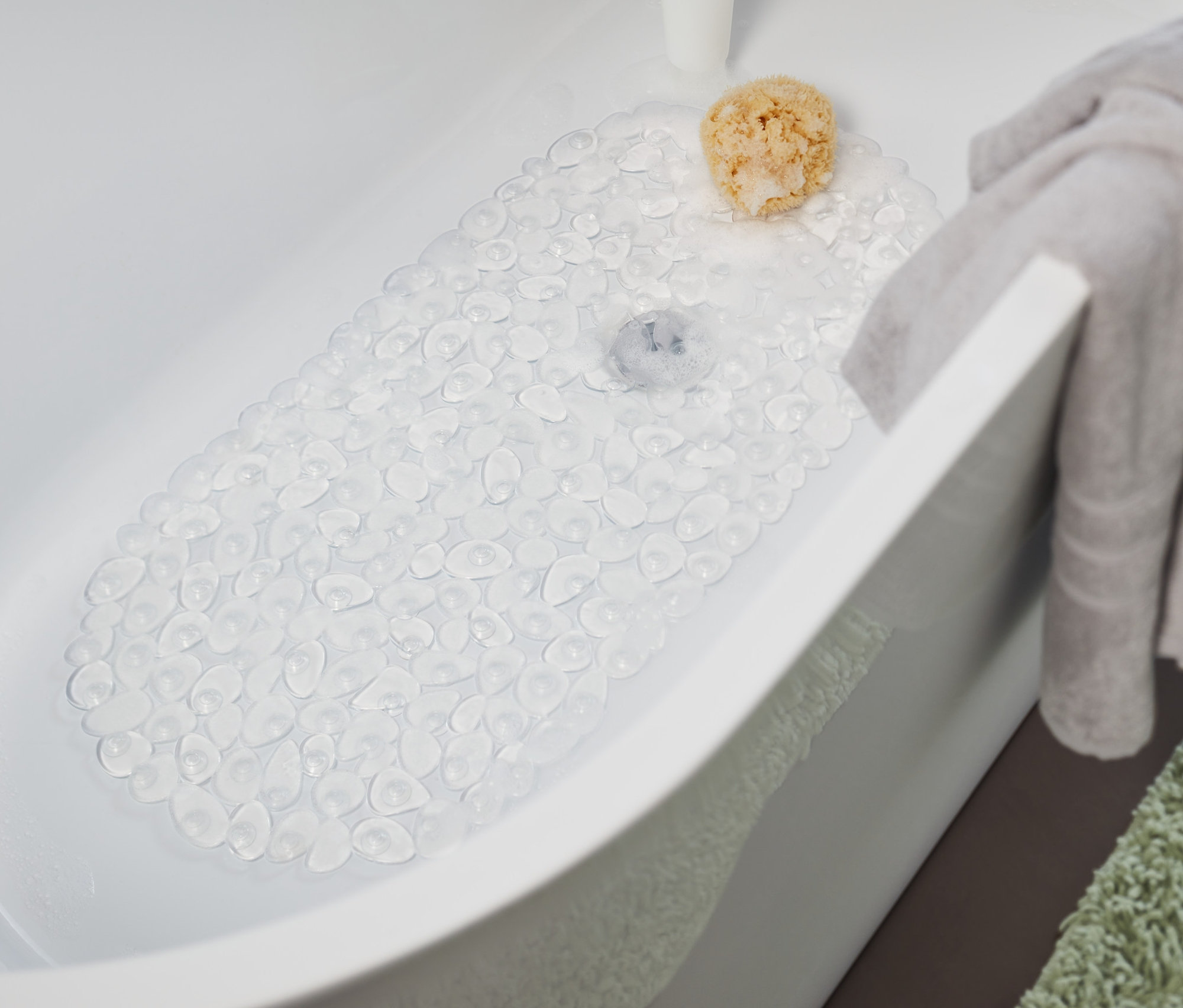 In der Badewanne liegt eine transparente Badewanneneinlage. Ein graues Handtuch hängt am Rand der Badewanne und ein grüner Teppich liegt neben der Badewanne.