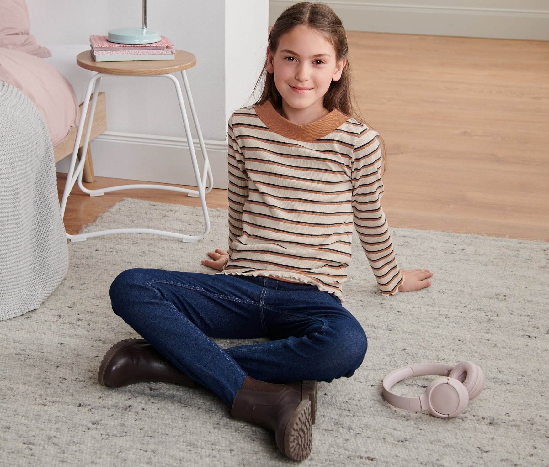 Mädchen sitzt in einem geringelten Rippshirt, Jeans und Stiefeln auf dem Teppich. Neben ihr liegen Kopfhörer.