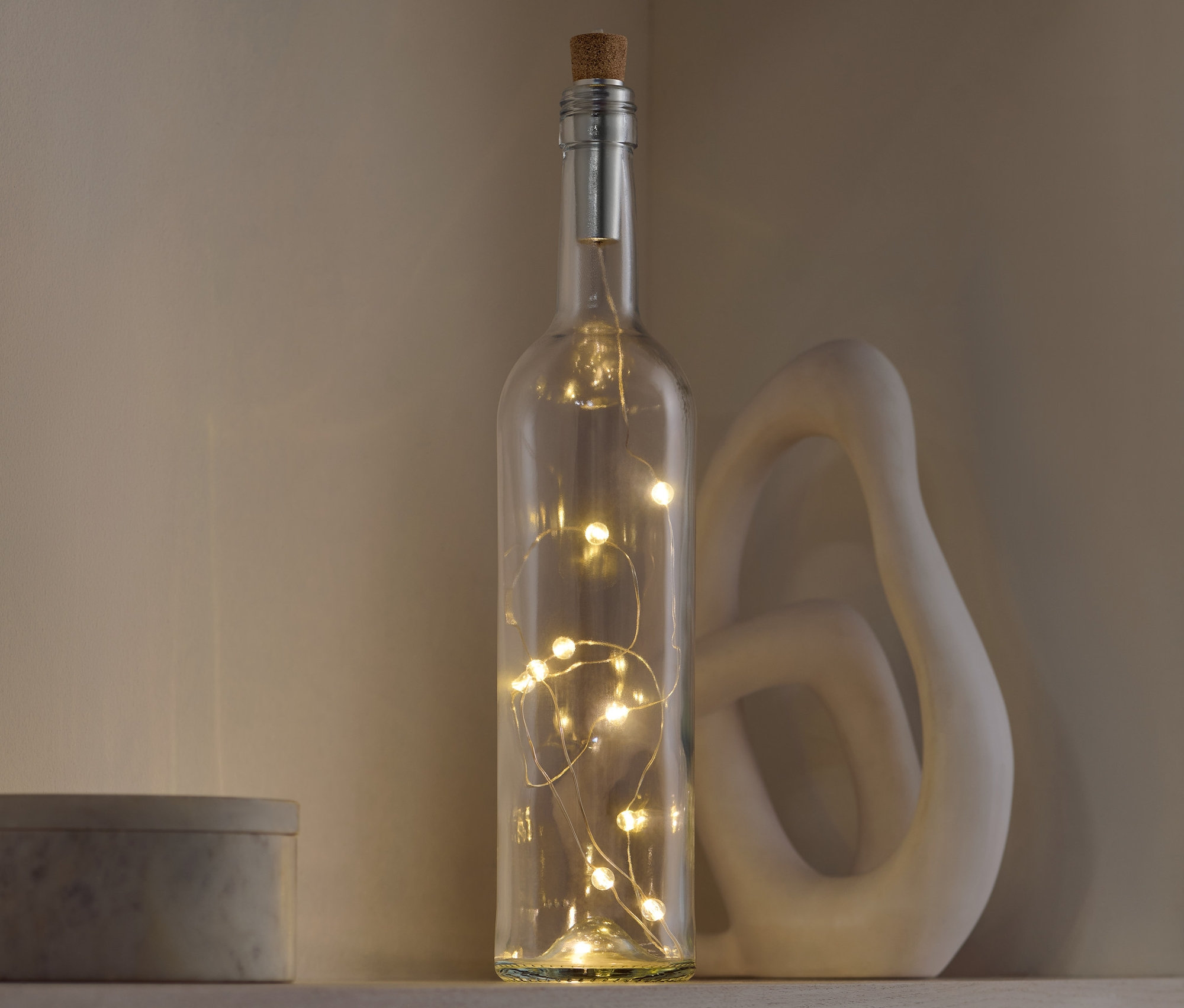 Dekorative Glasflasche mit Korkverschluss und Lichtern steht auf einem Regal neben einer dekorativen Skulptur und einer grauen Schachtel.