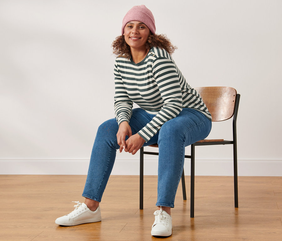 Eine Frau sitzt auf einem Stuhl und trägt ein gestreiftes Langarmshirt, eine Strickmütze mit Wolle, rosé, und eine Slimfit-Jeans.
