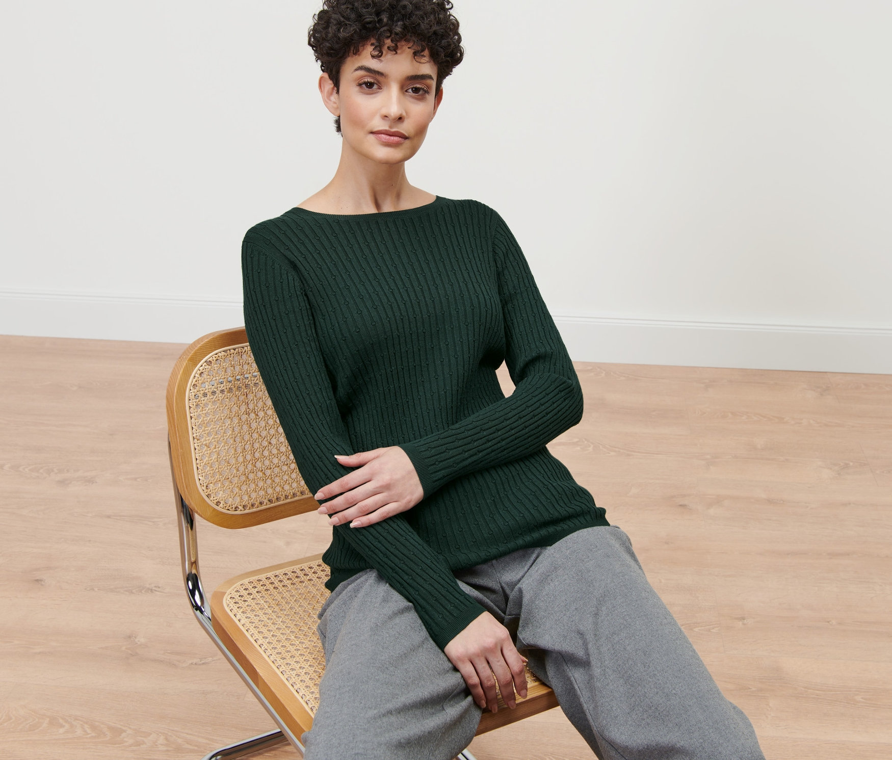 Frau sitzt auf einem Stuhl und trägt einen grünen Zopfstrickpullover.