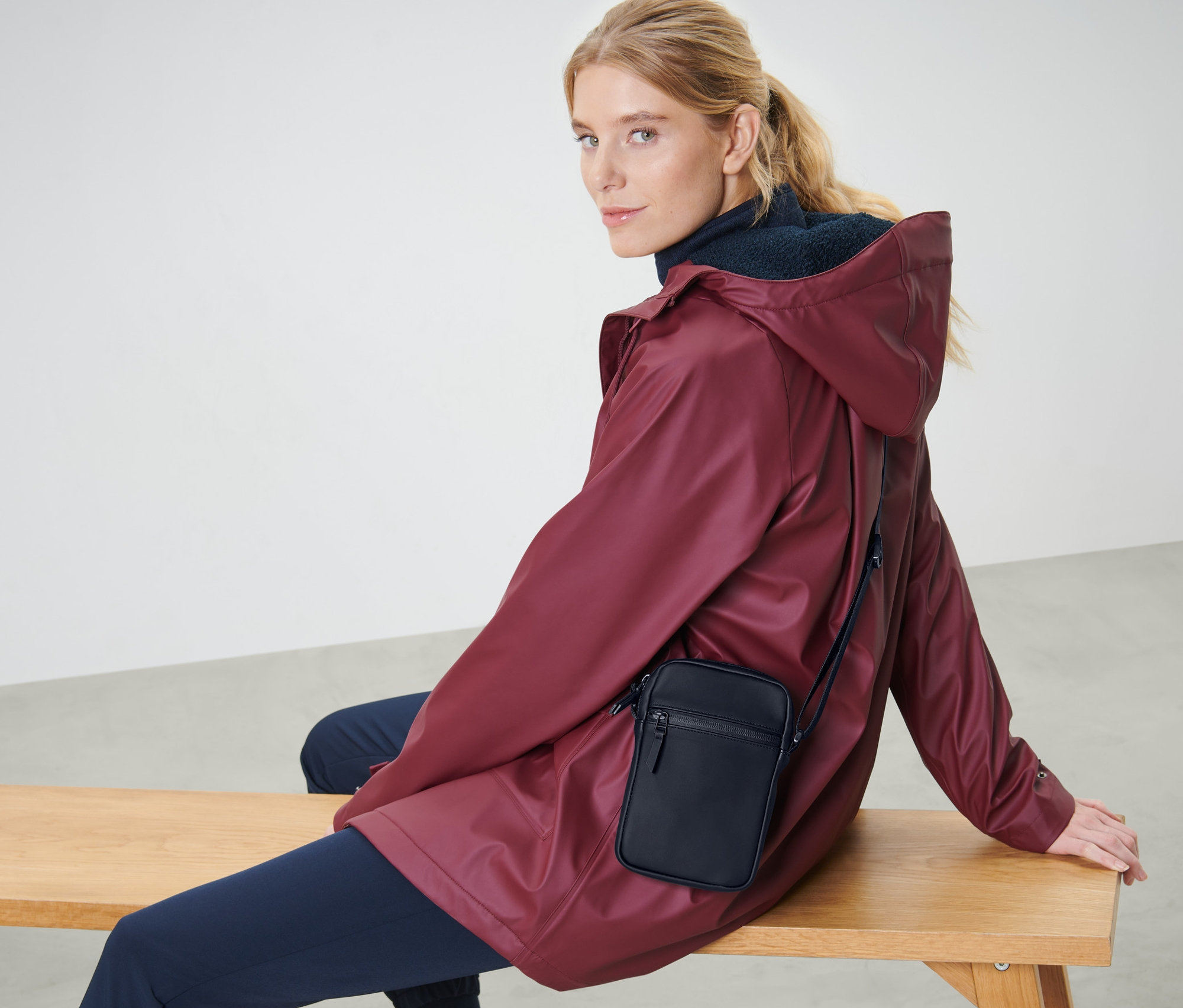 Blonde Frau sitzt auf einer Bank mit einer bordeauxroten Jacke und einer schwarzen Regen-Umhängetasche.