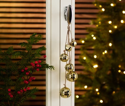 Glöckchen-Anhänger hängt an einer Tür, neben einer Vase mit Zweigen und im Hintergrund ein Weihnachtsbaum.