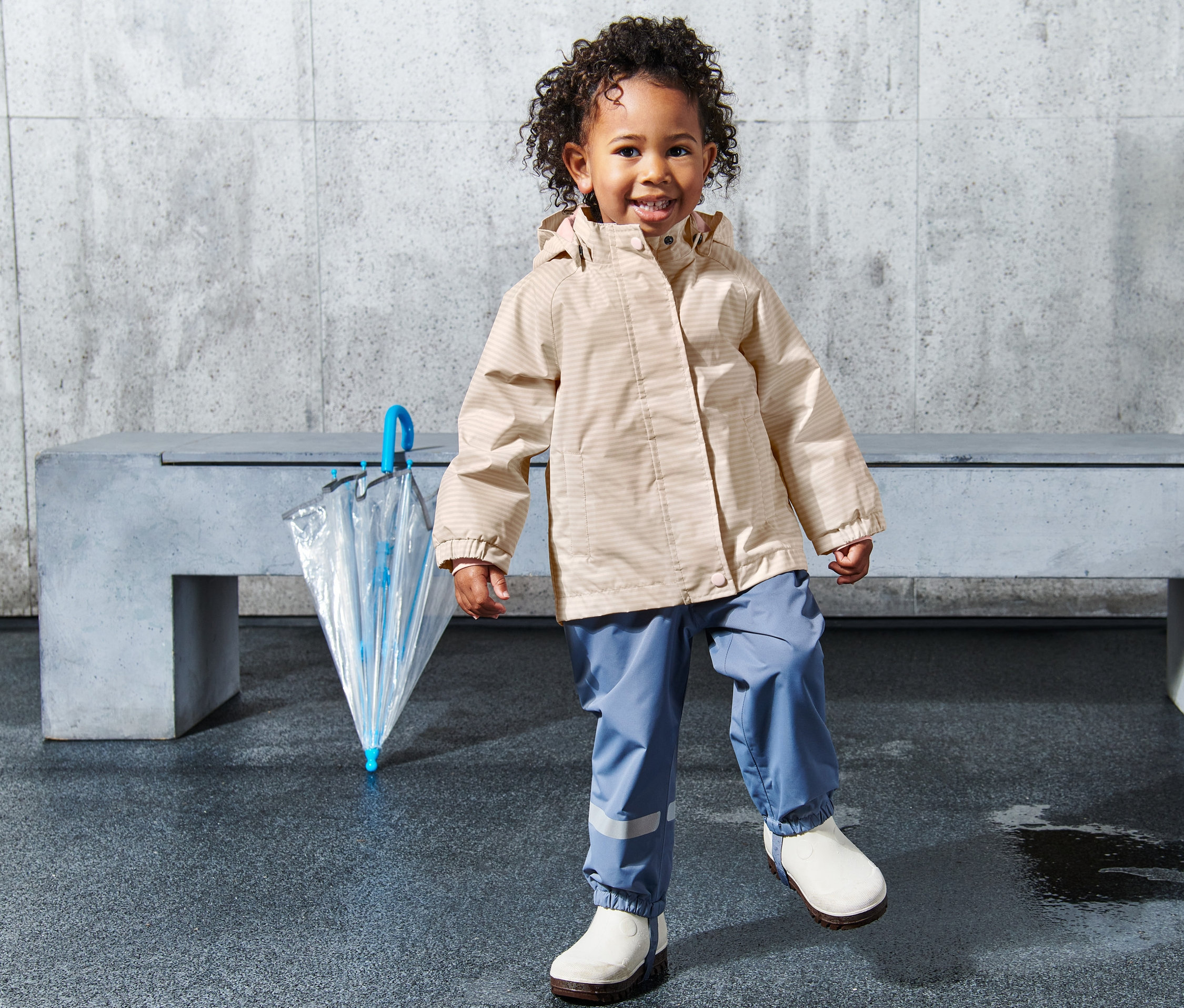 Ein Kleinkind steht auf nassem Boden neben einem durchsichtigen Regenschirm. Es trägt eine gestreifte Kleinkinder-Regenjacke und blaue Hosen.