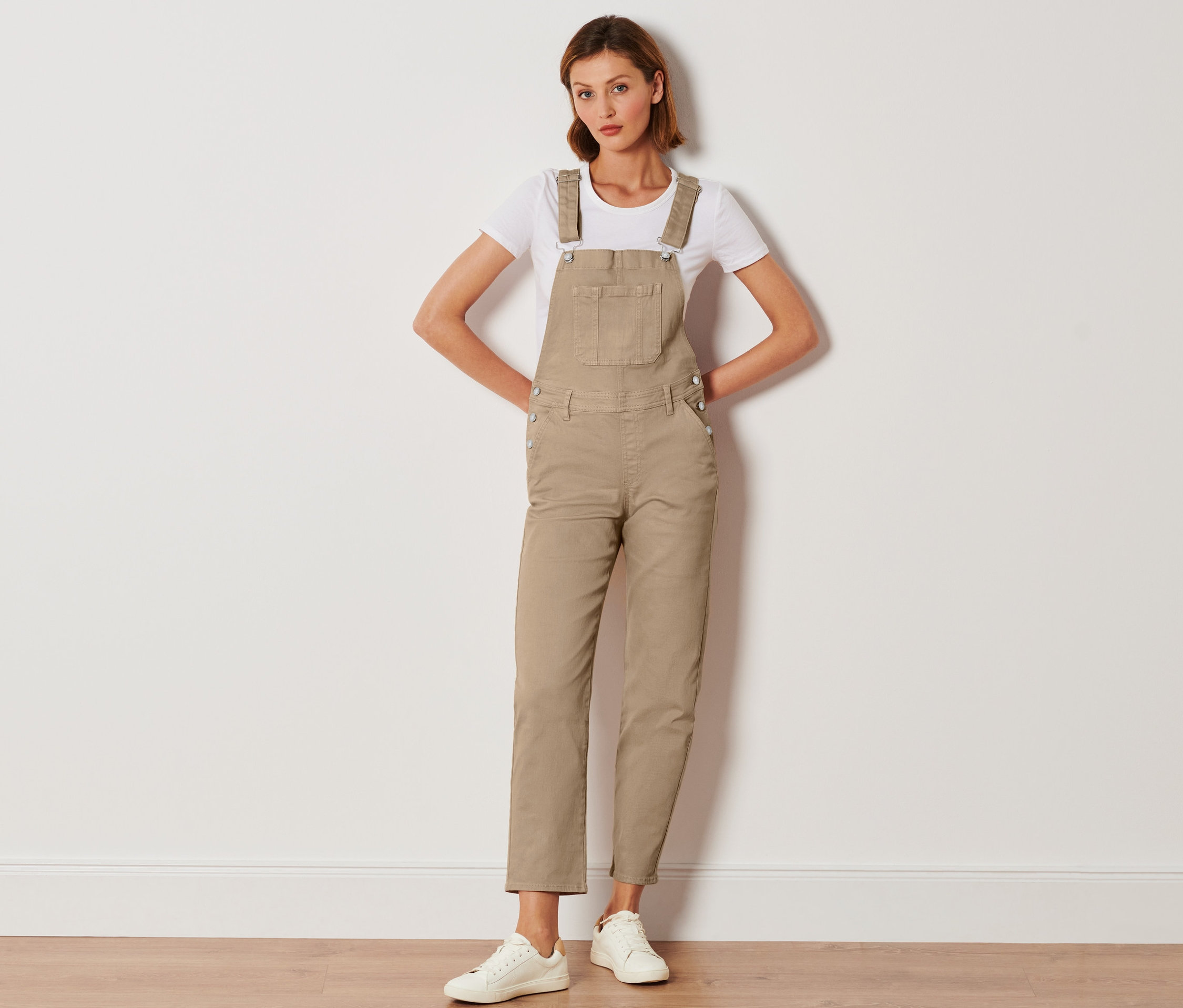 Frau posiert in beiger Latzhose, weißem T-Shirt und weißen Turnschuhen.