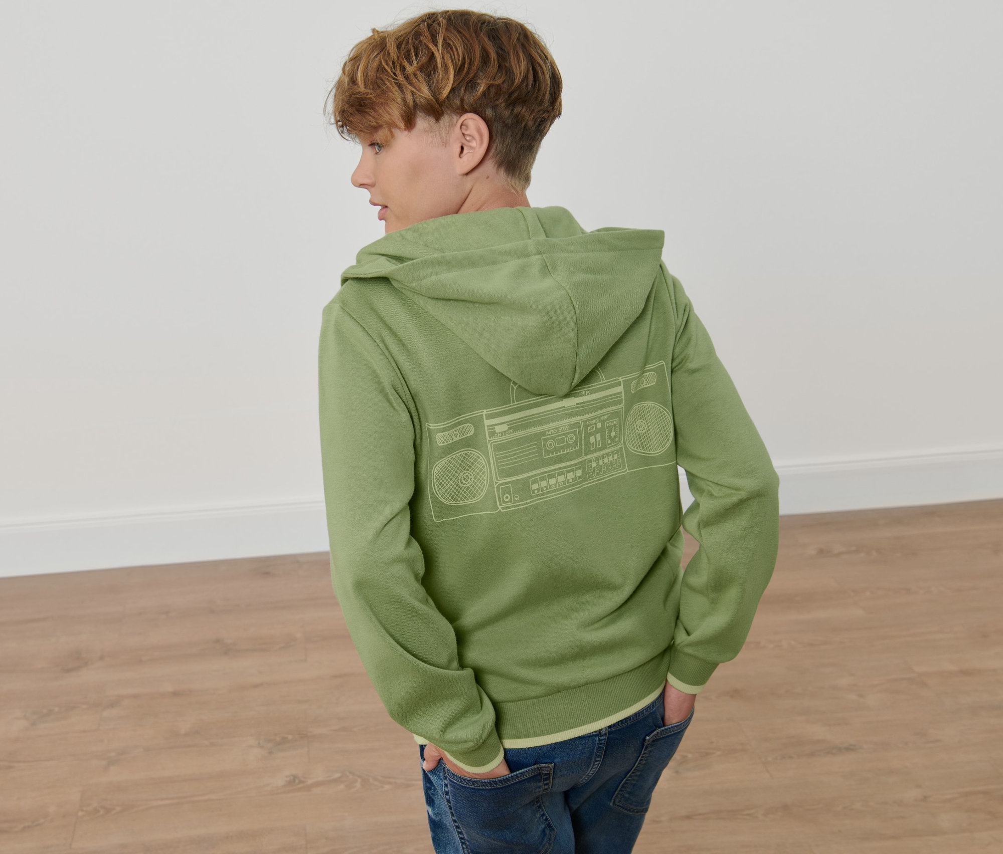 Junge von hinten trägt eine grüne Kinder-Sweatjacke mit Kapuze und Boombox-Aufdruck und Jeans.