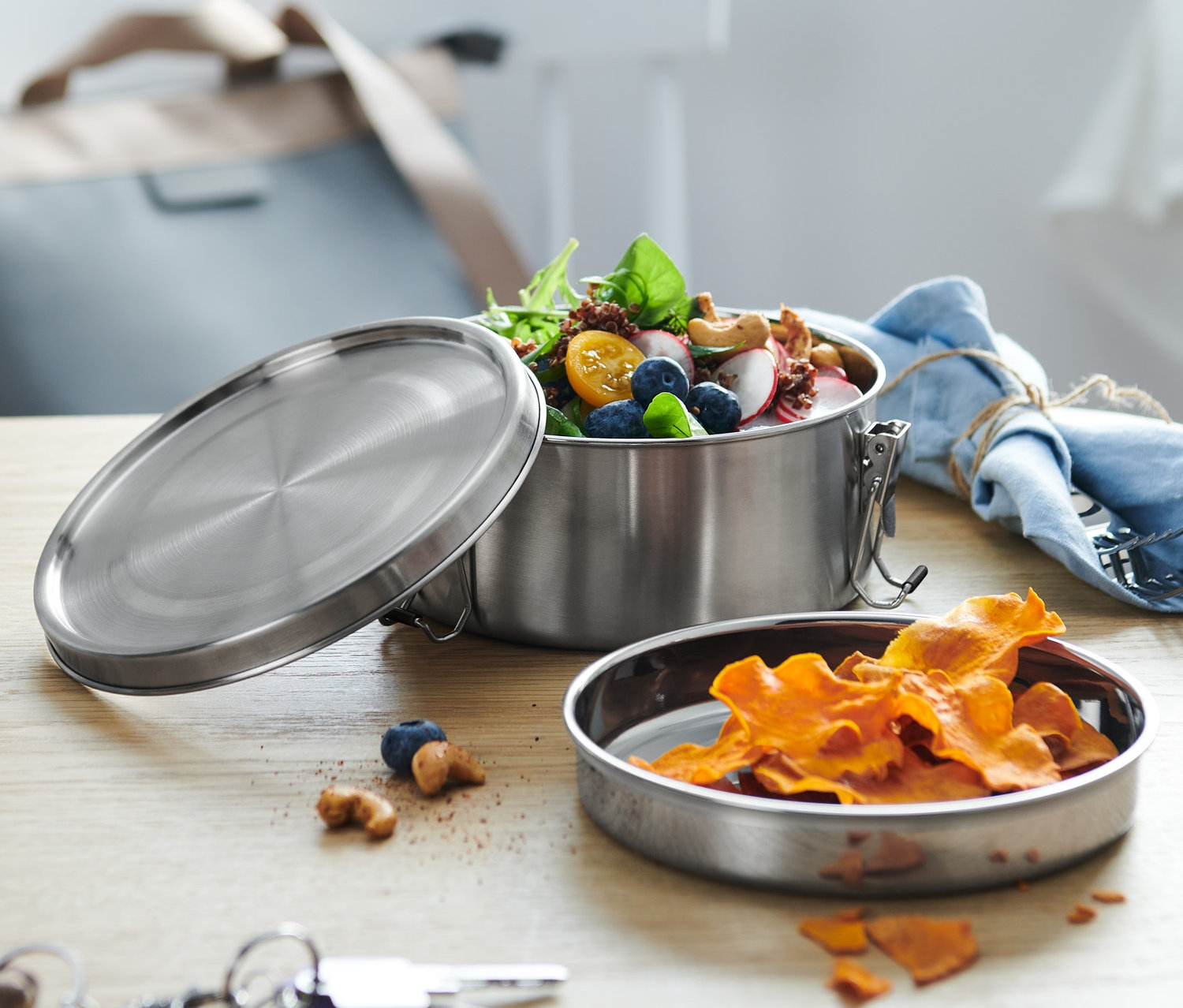 Eine Edelstahl-Lunchbox mit Salatfüllung, daneben liegen der Deckel und ein Teller mit Chips.
