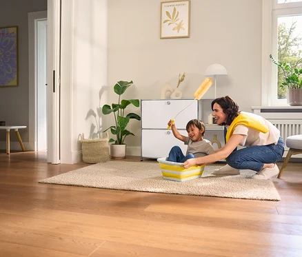 Eine Frau schiebt ein Kind in einem faltbaren Wäschekorb über einen Teppich. Im Hintergrund befinden sich ein Sideboard Metall »CN3« mit 2 Klappenfächern, weiß und eine Heizkörperbürste.