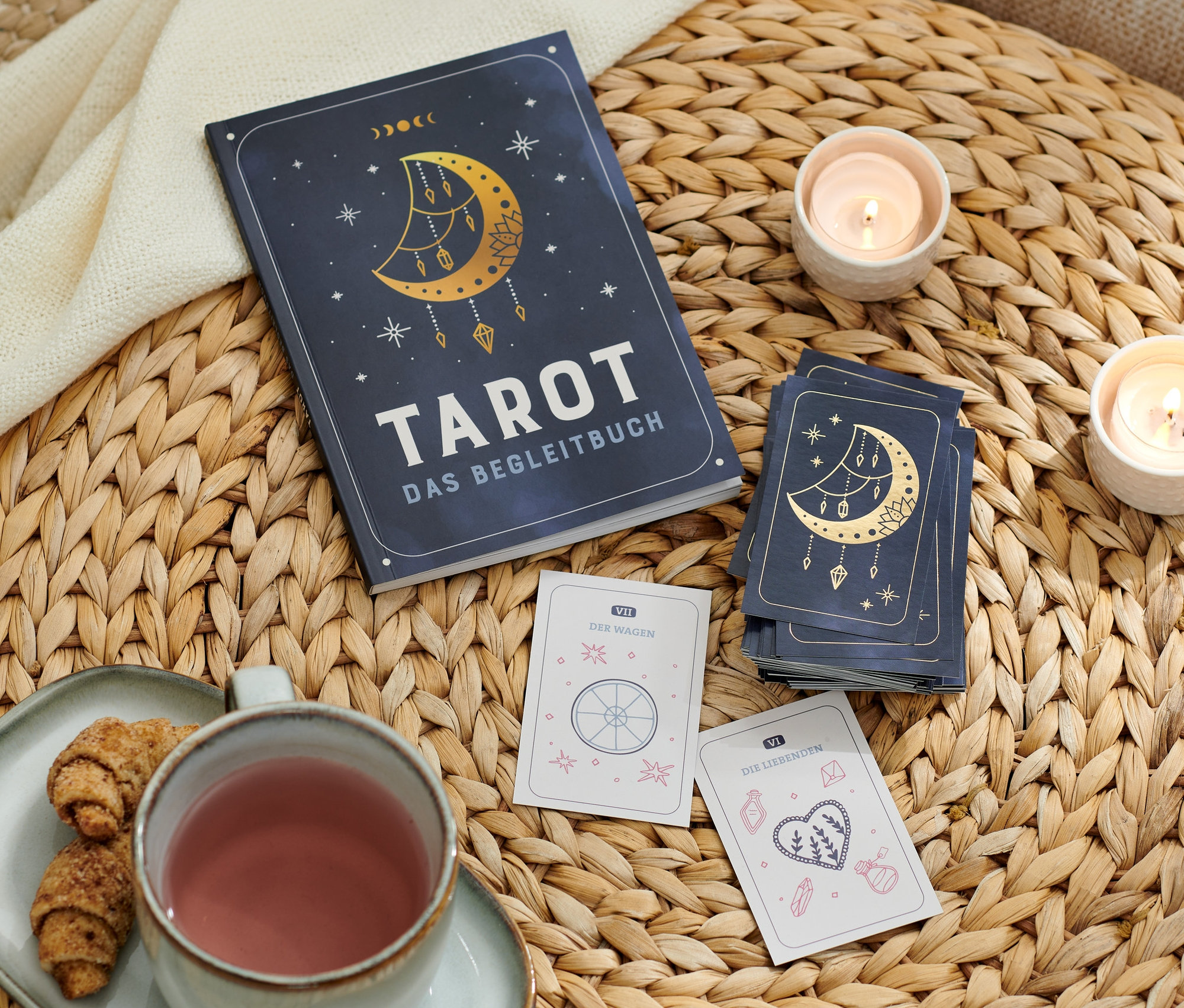 Auf einem Korbtisch liegen Tarot – das Einsteiger-Set mit Karten und Begleitbuch, Tarotkarten, zwei Kerzen, eine Tasse und ein Teller mit Croissants.