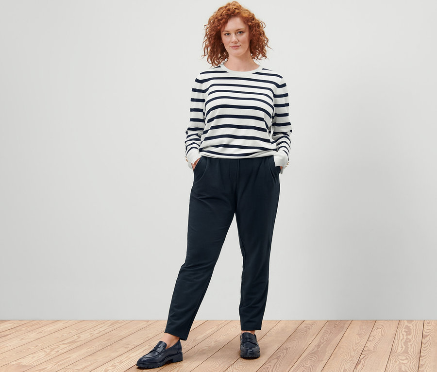 Frau steht mit Händen in den Taschen einer Joggpants und trägt einen gestreiften Feinstrickpullover, weiß.