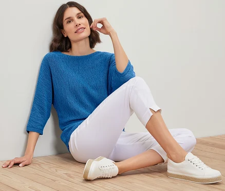 Frau sitzt auf Holzboden und trägt einen blauen sommerlichen Strickpullover aus Bändchengarn, weiße Hose und Turnschuhe.