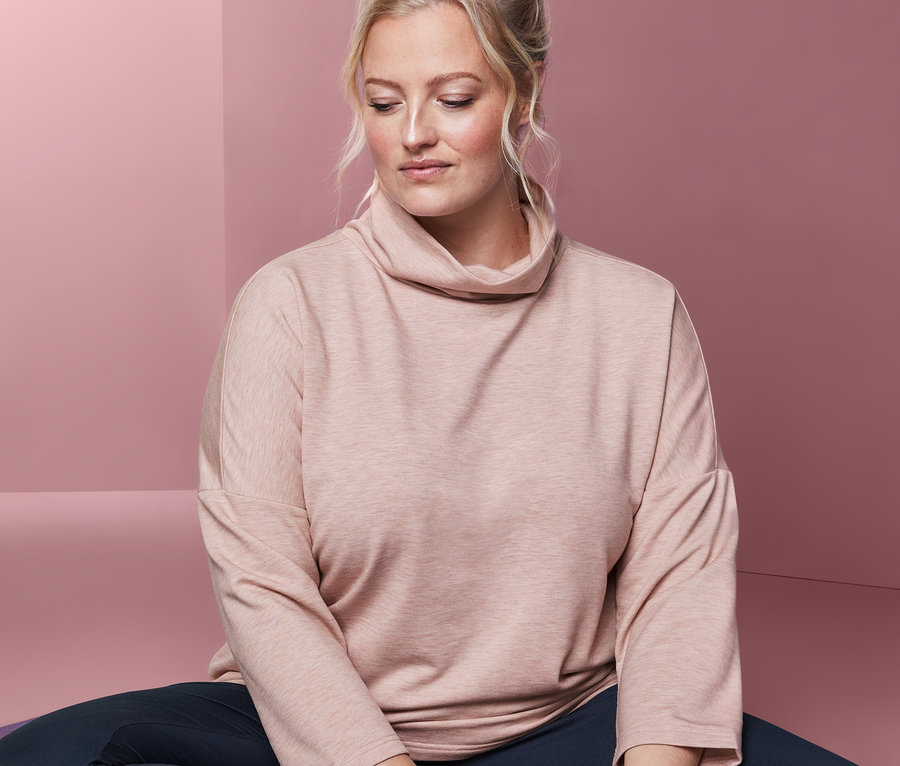 Blonde Frau sitzt mit gekreuzten Beinen auf einer lila Yogamatte und trägt einen Sweatpullover.