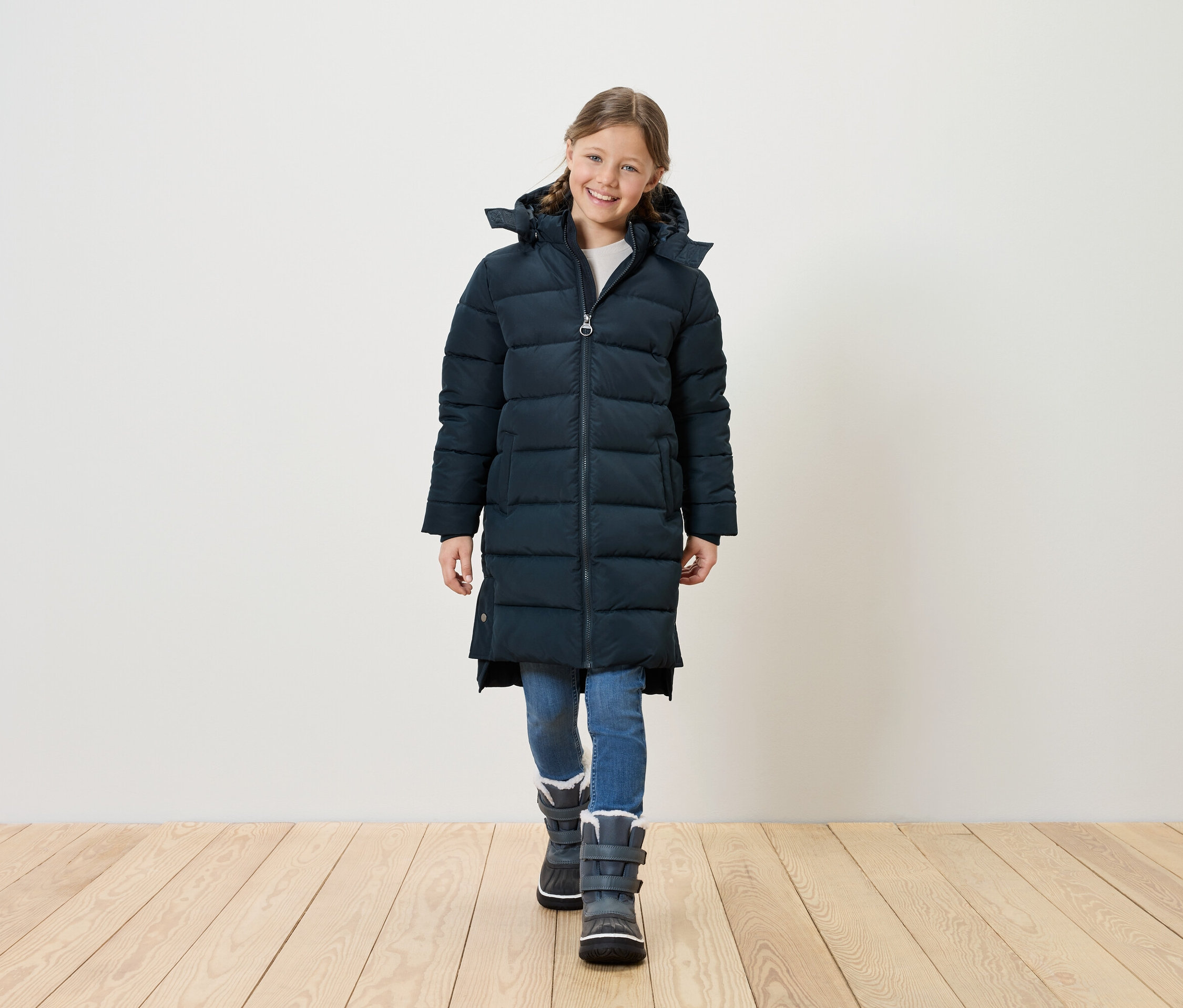 Lächelndes Mädchen in langer, gesteppter, marineblauer Jacke, blauen Jeans und grauen Winterstiefeln steht auf einem Holzboden vor einer weißen Wand.