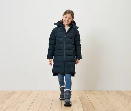 Lächelndes Mädchen in langer, gesteppter, marineblauer Jacke, blauen Jeans und grauen Winterstiefeln steht auf einem Holzboden vor einer weißen Wand.