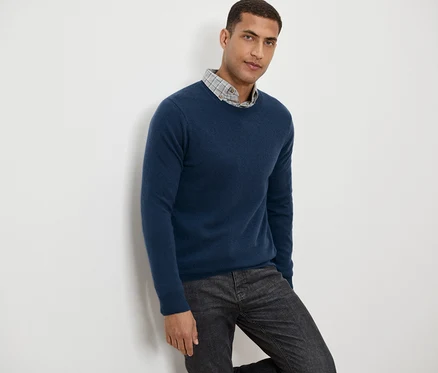 Mann lehnt an einer Wand, trägt einen blauen Pullover und eine graue gemusterte Hose.