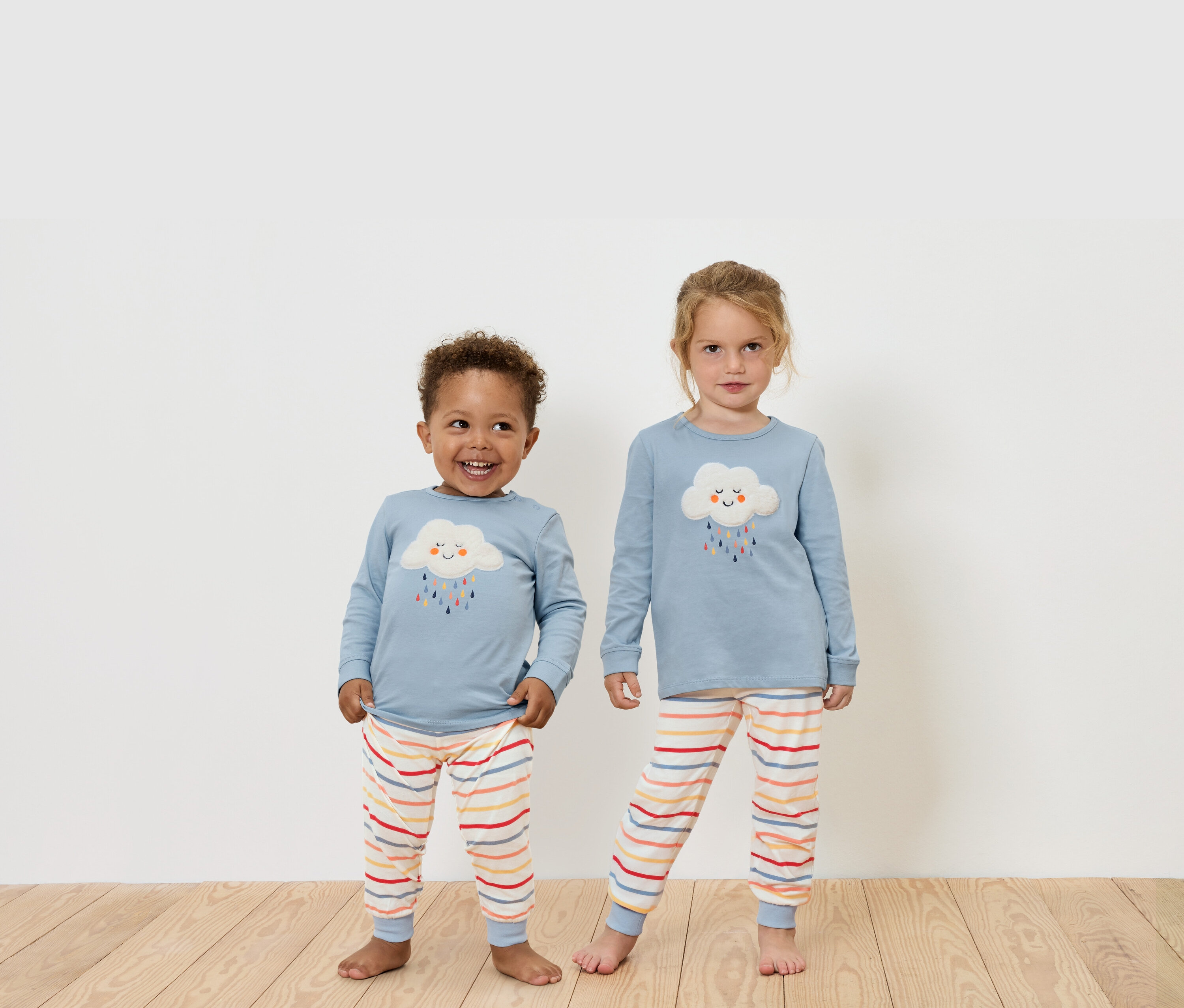 Zwei Kinder stehen auf einem Holzboden und tragen blaue Langarmshirts und gestreifte Hosen.