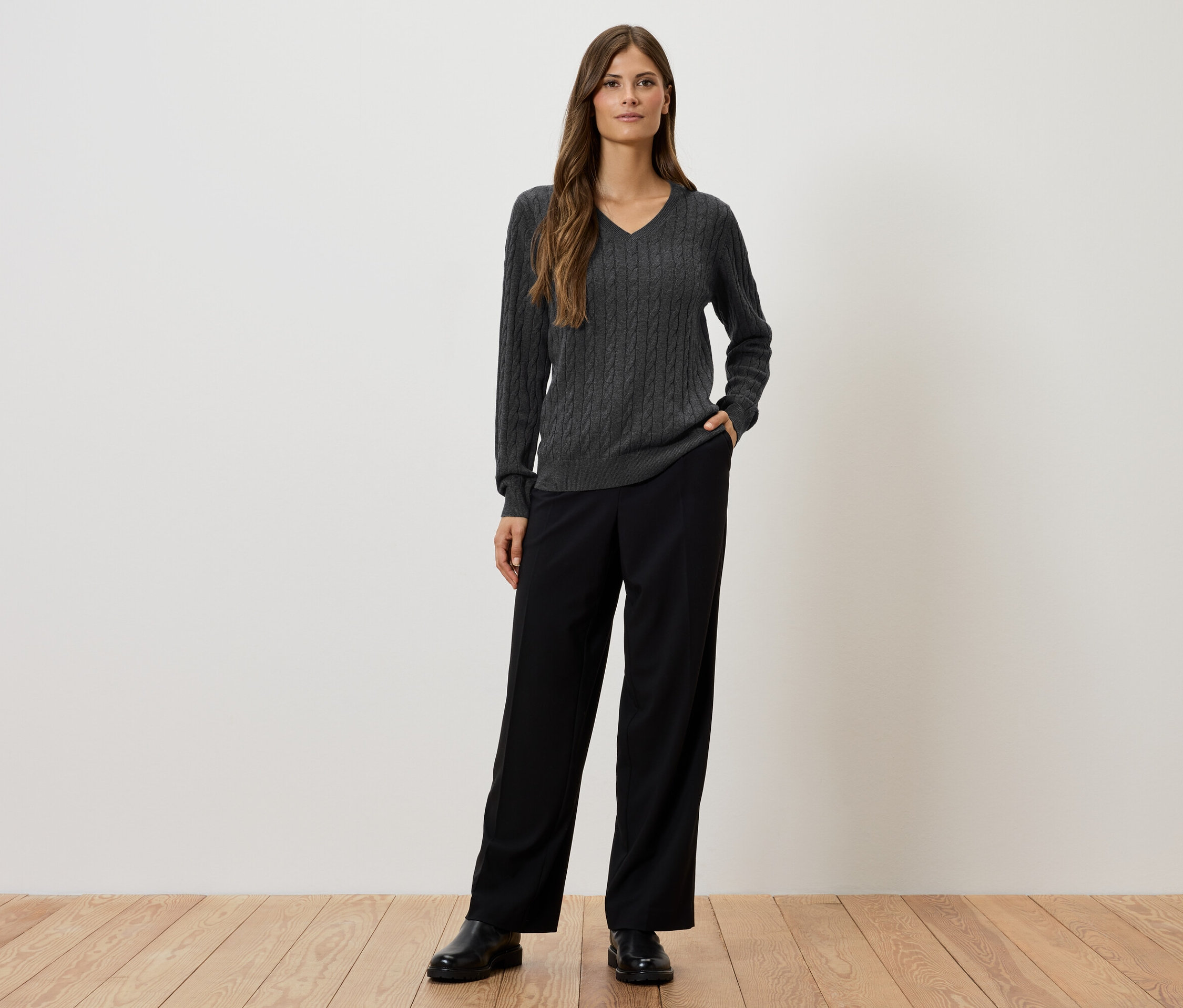 Eine Frau steht vor einer weißen Wand und trägt einen grauen Zopfmusterpullover, eine schwarze Hose und schwarze Schuhe.