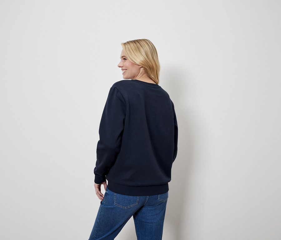 Blonde Frau blickt über die Schulter, trägt einen blauen Pullover und Jeans.