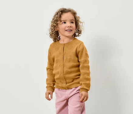 Mädchen mit lockigem Haar trägt eine braune Strickjacke mit Herzen und eine rosa Hose.